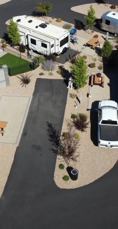 Aerial view of an RV park with two campers parked on gravel sites. A white truck is beside one camper, and picnic tables surrounded by trees are visible.
