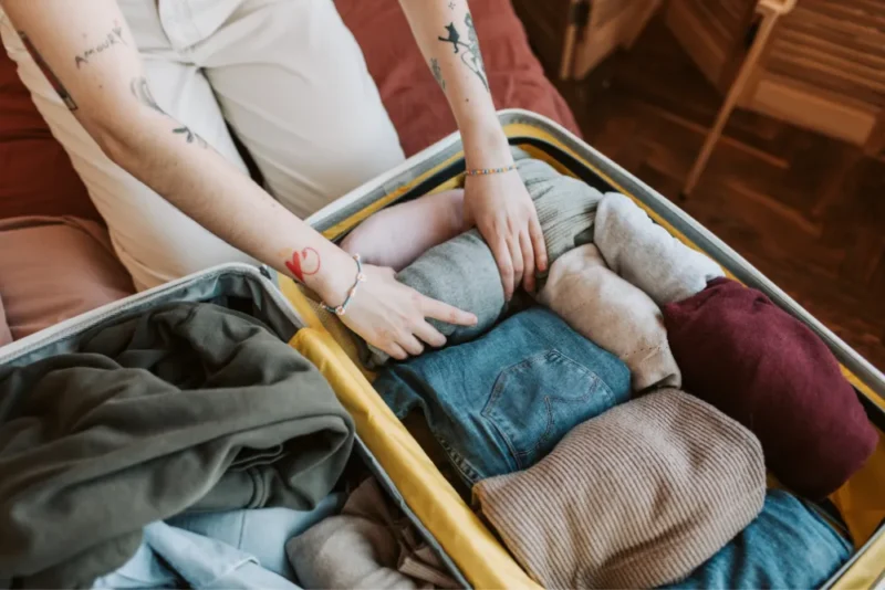 Person with tattoos packs a suitcase with neatly folded clothes, including sweaters and jeans, conveying a sense of organization and travel preparation.