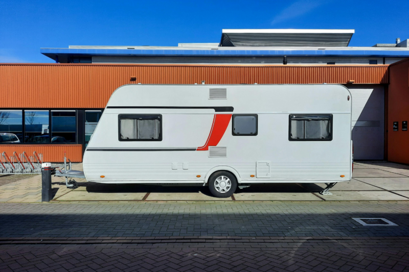 A white caravan with a red stripe is parked by an orange corrugated building under a clear blue sky, conveying a sense of travel and adventure.