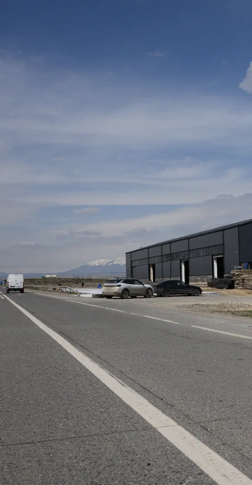 Roadside view with a modern warehouse and parked cars under a clear blue sky. Snow-capped mountains are visible in the background, conveying a peaceful tone.