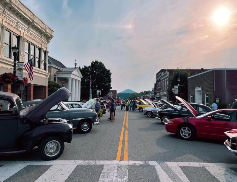 Downtown classic car show on a sunny historic street, with vintage vehicles, people, American flags, and distant blue mountains.