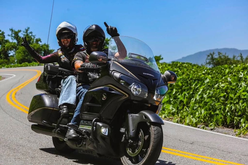 a couple on a motorcycle on the back of the dragon, throwing up a peace sign at the camera as they ride