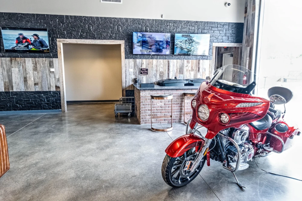 Bright motorcycle showroom with a polished red touring motorcycle in the foreground, TVs and a reception desk behind it; clean, welcoming atmosphere.