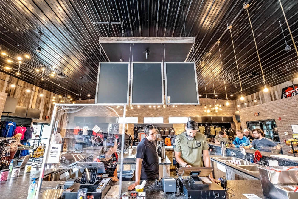 Busy modern café with baristas preparing drinks behind the counter, customers nearby, and a warm industrial interior with hanging lights.